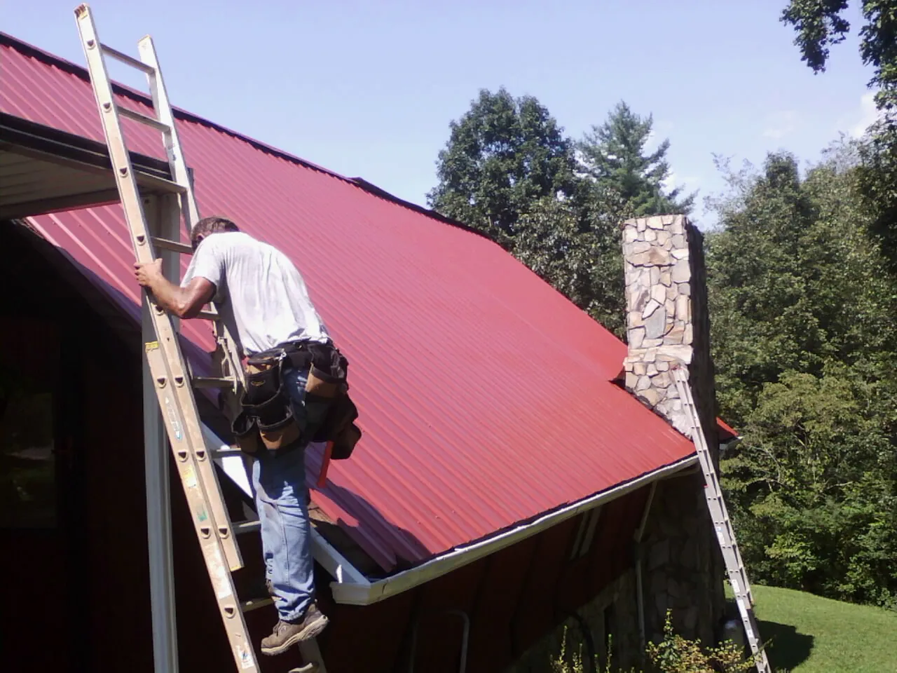 Roofer on a ladder beside a red metal roof and stone chimney — Soffit & Fascia Repair by Roofing Brothers in New Millport